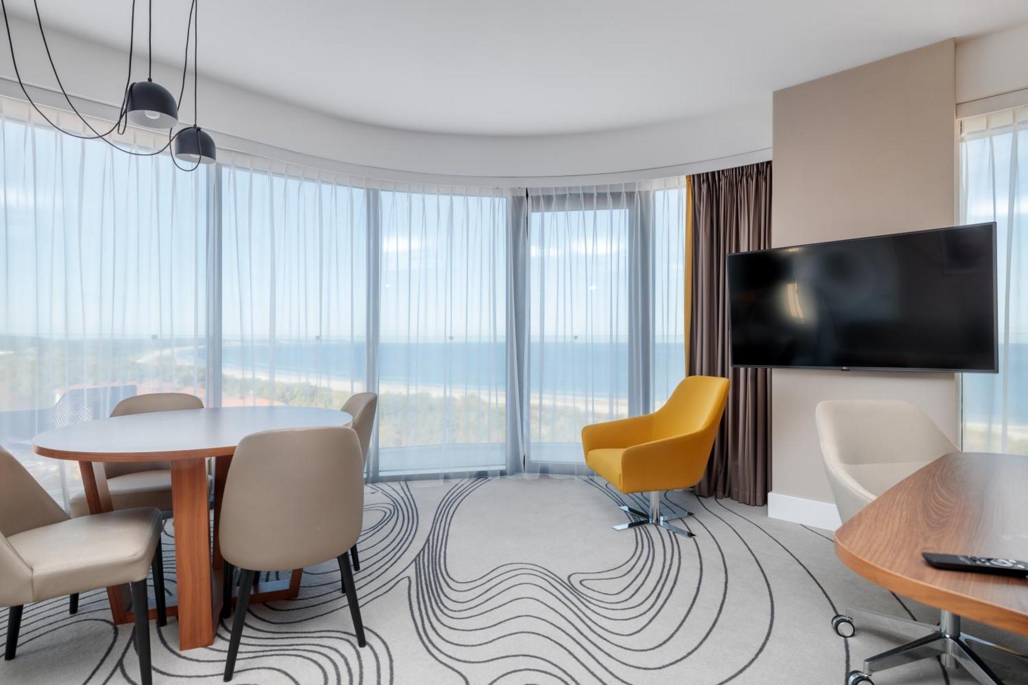 Radisson Blu Resort Świnoujście Εξωτερικό φωτογραφία The image shows a modern hotel room or suite with a bright, airy atmosphere. There are large windows that allow natural light to fill the space, providing a view of the ocean. The decor includes a round dining table with several chairs, and a stylish