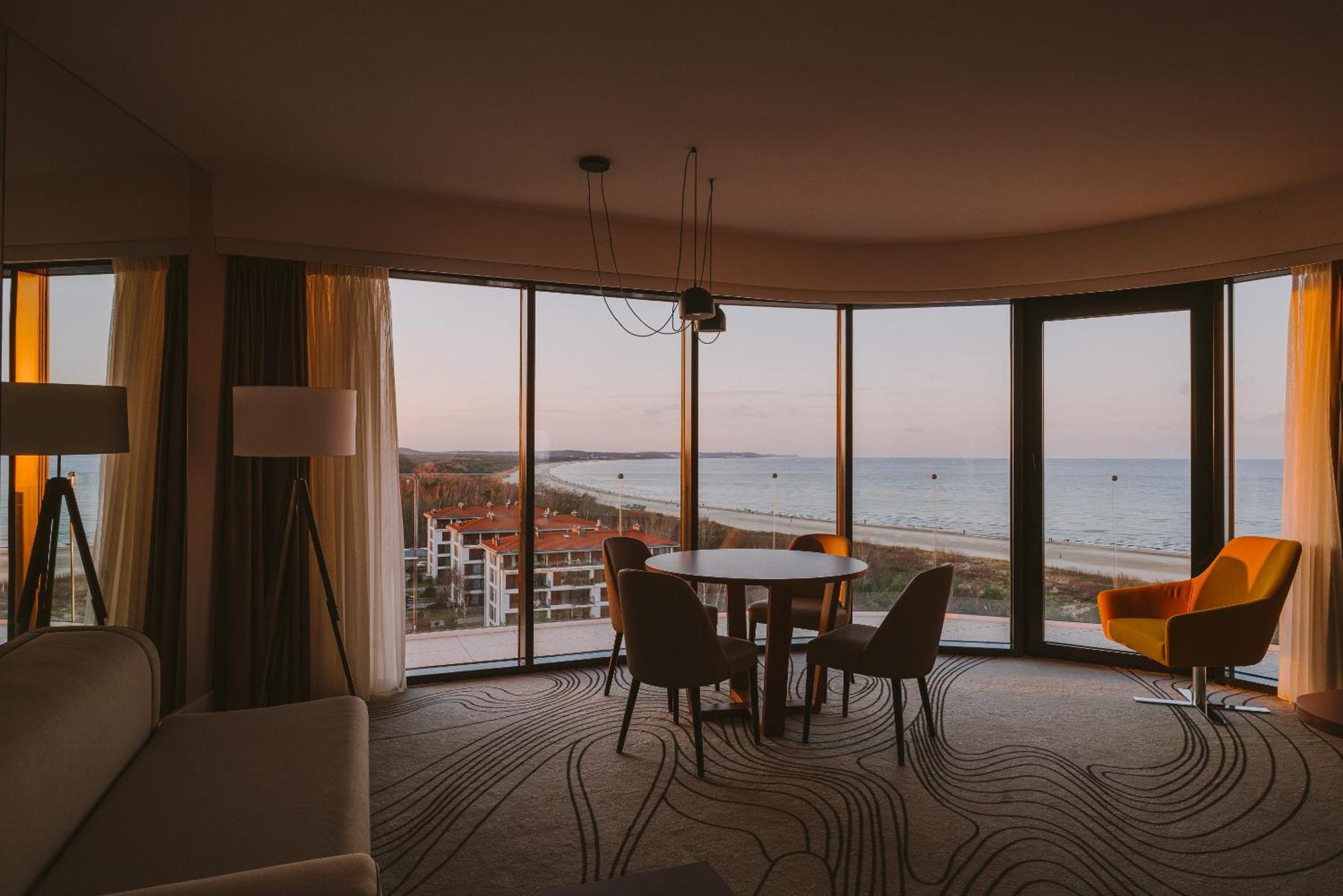 Radisson Blu Resort Świnoujście Εξωτερικό φωτογραφία The photo shows a modern and stylish interior of a room featuring large windows that provide a view of the beach and ocean. The room is furnished with a round table and chairs, along with a cozy sofa and a bright yellow armchair. The decor has a cont