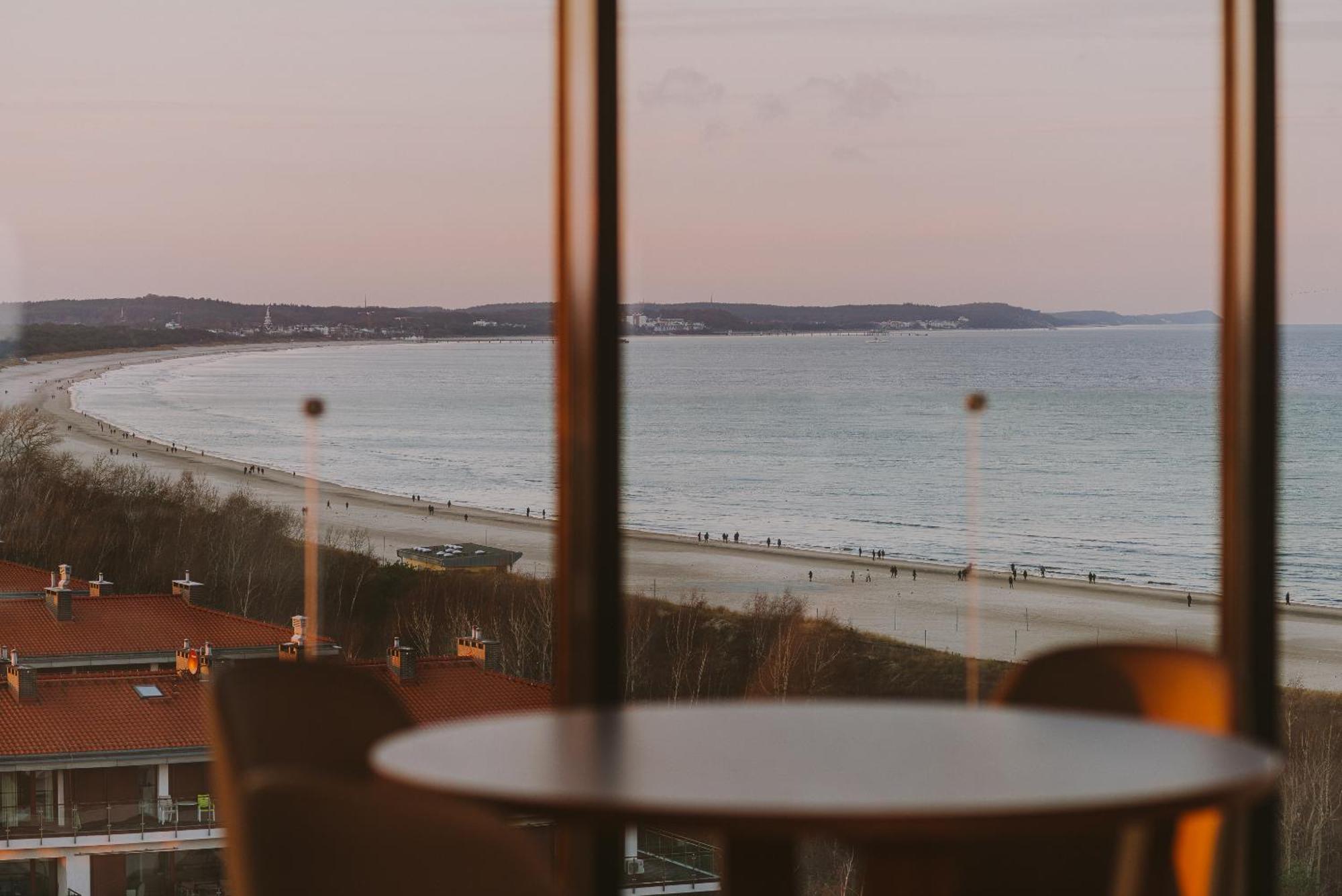 Radisson Blu Resort Świnoujście Εξωτερικό φωτογραφία The photo shows a picturesque view of a beach scene from indoors. In the foreground, there is a round table with some chairs around it, suggesting a cozy interior space. Through a large window, you can see the beach with gentle waves hitting the shor
