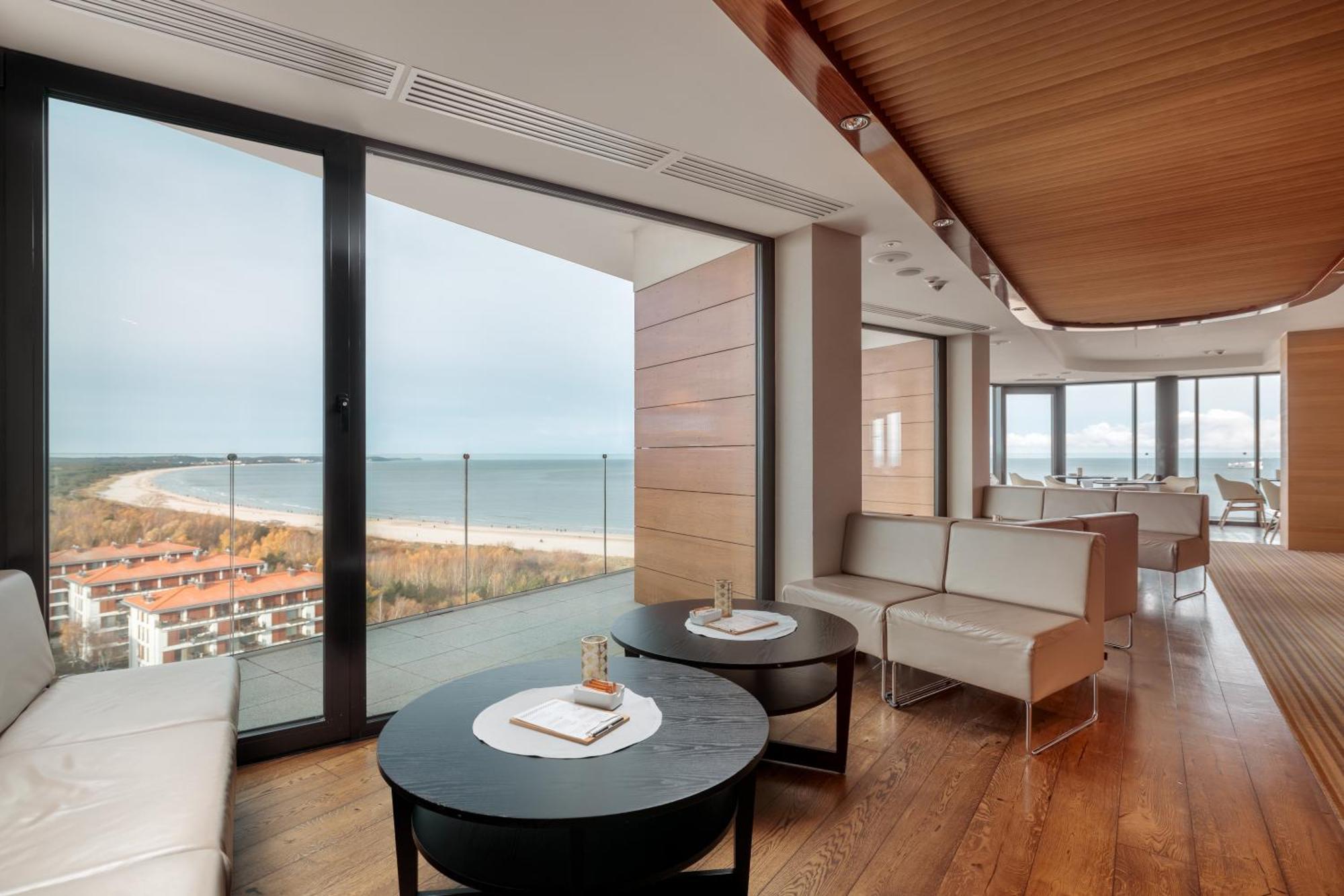 Radisson Blu Resort Świnoujście Εξωτερικό φωτογραφία The photo shows a modern, elegantly designed interior space featuring large windows that offer a panoramic view of the ocean and coastline. The flooring is wooden, providing a warm atmosphere. There are two round black tables in the foreground with m