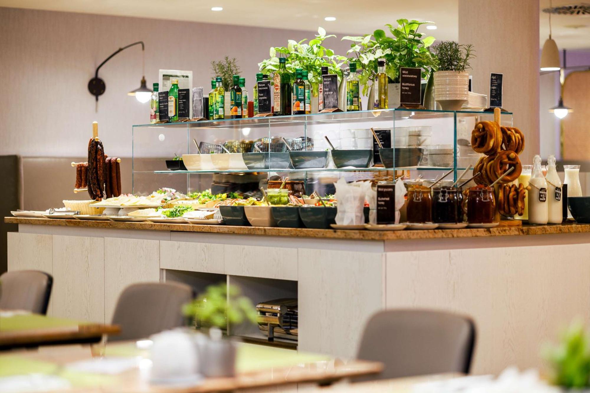 Radisson Blu Resort Świnoujście Εξωτερικό φωτογραφία The photo shows a well-organized buffet display in a restaurant setting. There is a glass counter filled with various dishes. On the upper shelf, there are several bottles of oil and herbs, indicating a focus on fresh and healthy ingredients. Below t