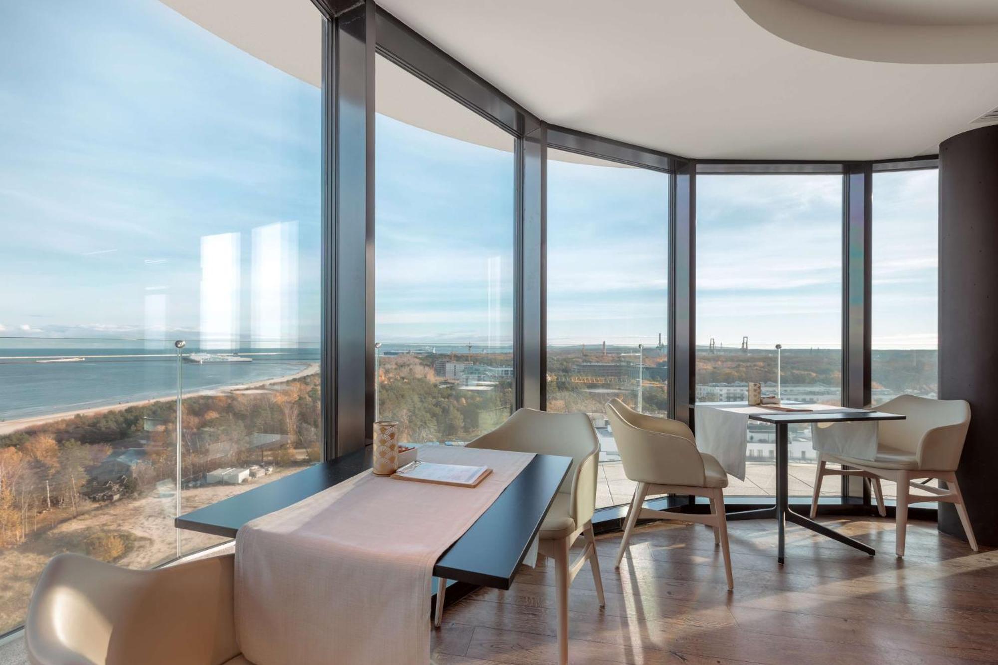 Radisson Blu Resort Świnoujście Εξωτερικό φωτογραφία The photo shows a modern dining area with large windows that offer a panoramic view of the landscape outside, including a body of water and possibly a shoreline. The design features sleek furniture, including a table with chairs arranged around it, a
