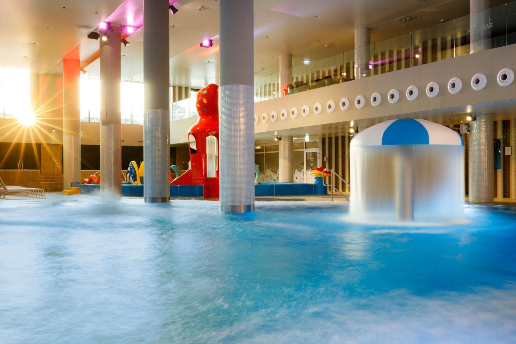 Radisson Blu Resort Świnoujście Εξωτερικό φωτογραφία The photo shows an indoor water park or pool area. There is a large body of water with playful features, including a water slide shaped like a red bear and a smaller structure with a blue and white canopy. The lighting is bright, suggesting a sunny a