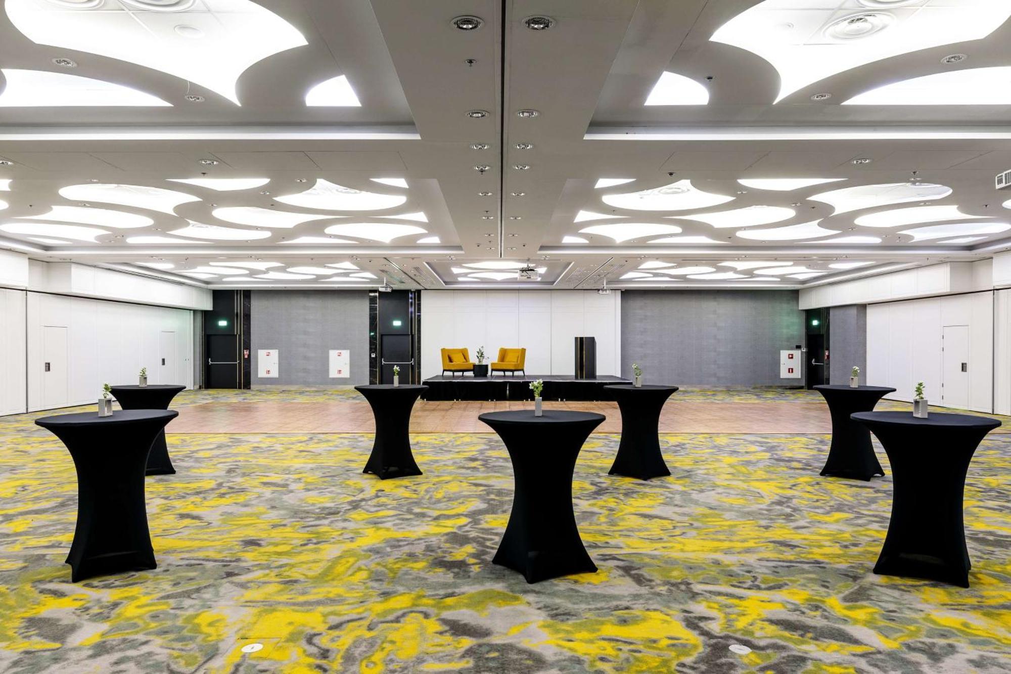 Radisson Blu Resort Świnoujście Εξωτερικό φωτογραφία The photo shows a spacious interior room set up for an event. The flooring features a patterned design in gray with yellow accents. There are several black tall tables scattered throughout the area, and in the background, there are two large yellow a