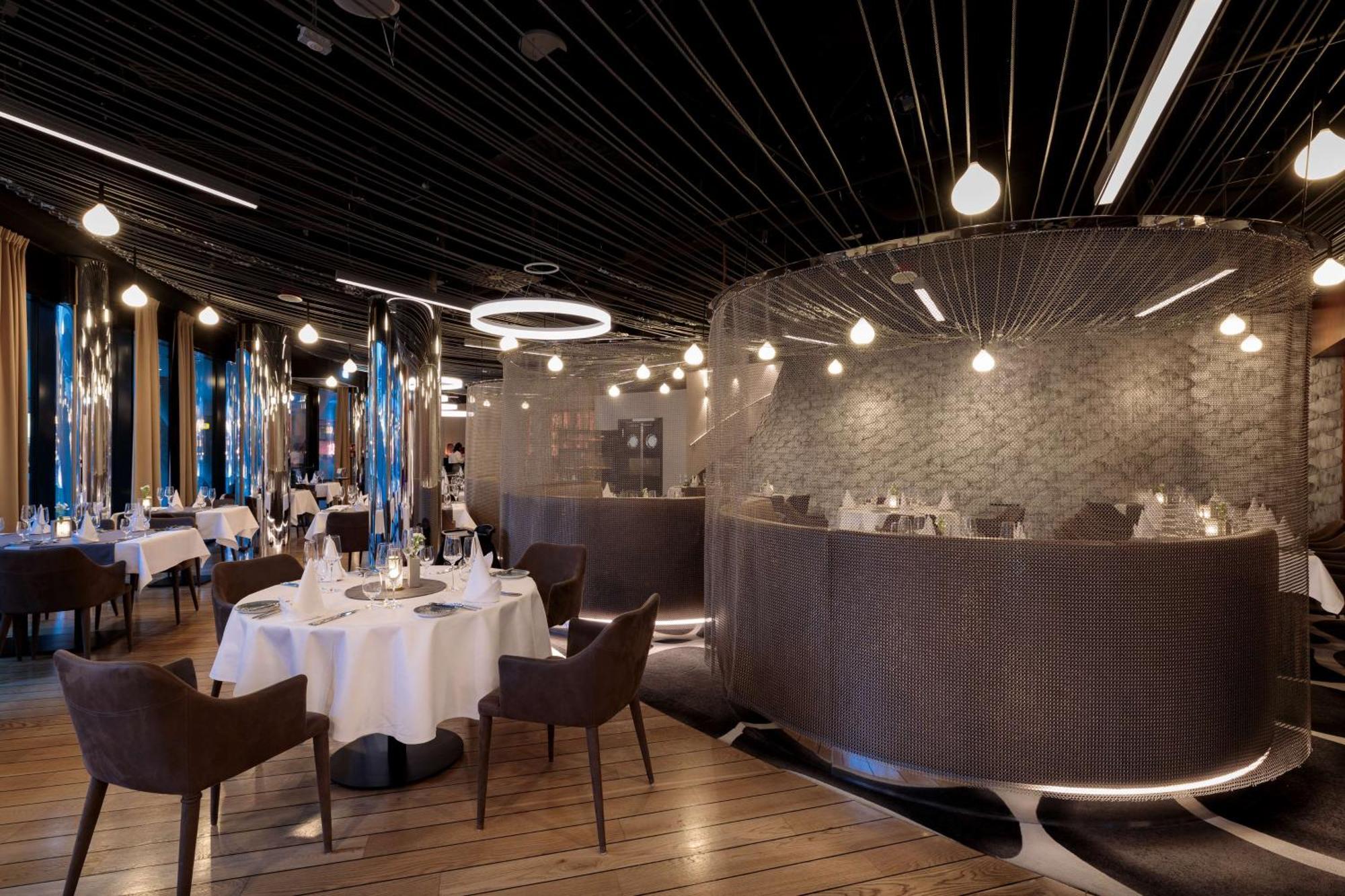 Radisson Blu Resort Świnoujście Εξωτερικό φωτογραφία The photo shows an elegant restaurant interior. It features polished wooden floors and contemporary decor. Round tables are set with white tablecloths and have chairs arranged around them. The lighting is modern, with multiple pendant lights hanging 