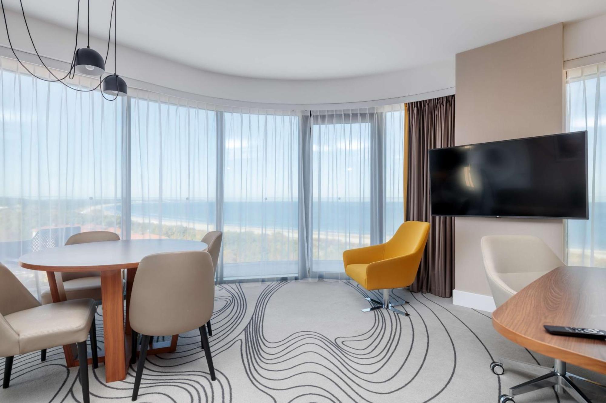Radisson Blu Resort Świnoujście Εξωτερικό φωτογραφία The photo shows a modern, stylish interior of a room with large windows that provide a panoramic view. The space features a round dining table surrounded by chairs, including a distinct yellow armchair. There is a flat-screen television mounted on th