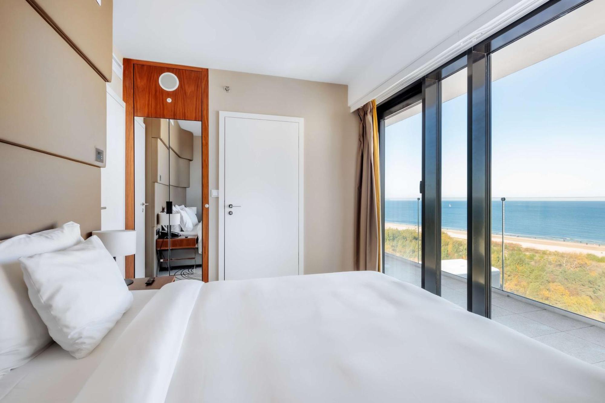 Radisson Blu Resort Świnoujście Εξωτερικό φωτογραφία The photo shows a modern hotel room with a bright and airy atmosphere. There is a large bed with white bedding positioned near a glass wall that offers a view of the ocean. The room features neutral-colored walls and wooden accents, enhancing its con