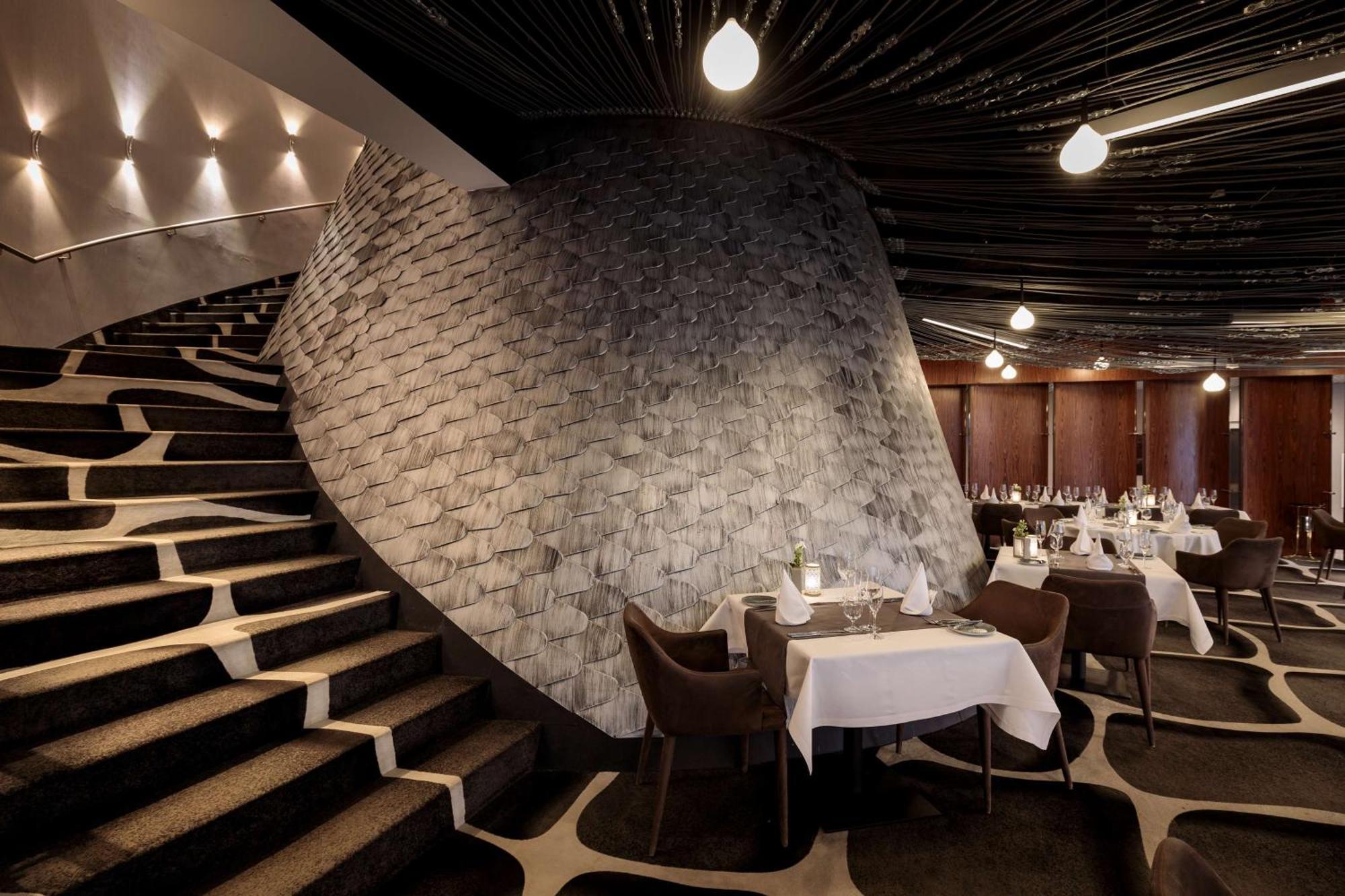 Radisson Blu Resort Świnoujście Εξωτερικό φωτογραφία The photo shows the interior of a stylish restaurant. In the foreground, there are elegantly set tables with white tablecloths and dark chairs. A unique, curving stone wall adds a distinct architectural feature, and a grand staircase with a textured 
