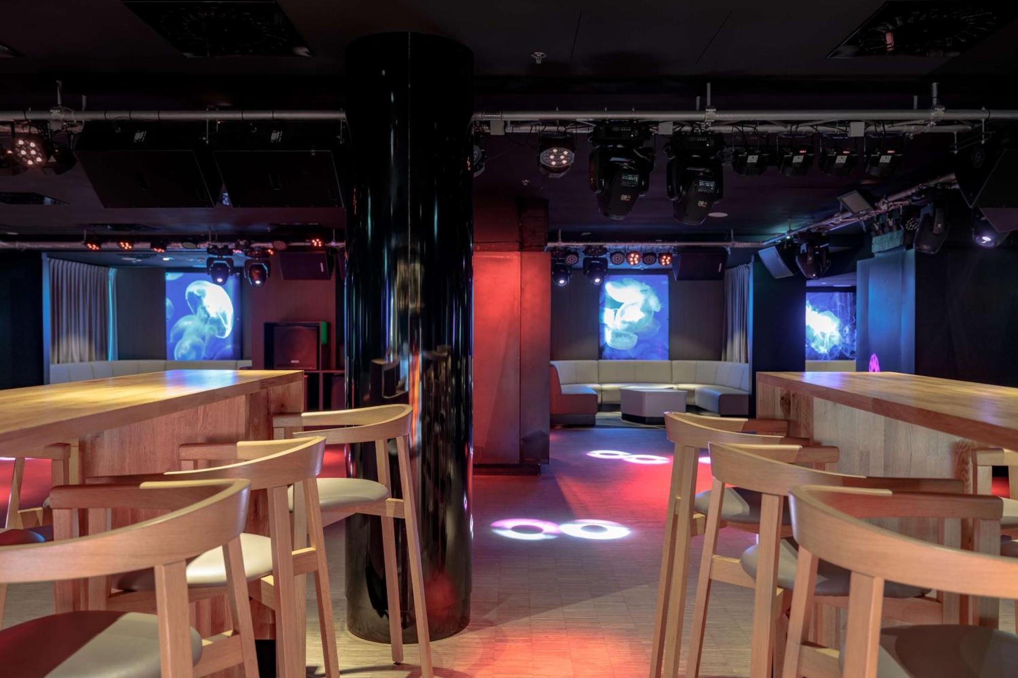 Radisson Blu Resort Świnoujście Εξωτερικό φωτογραφία The photo shows an interior of a modern venue, likely a bar or club. The space features a central black column and light-colored wooden bar stools arranged around a bar area. On the walls, there are large screens displaying vibrant visuals, possibly 